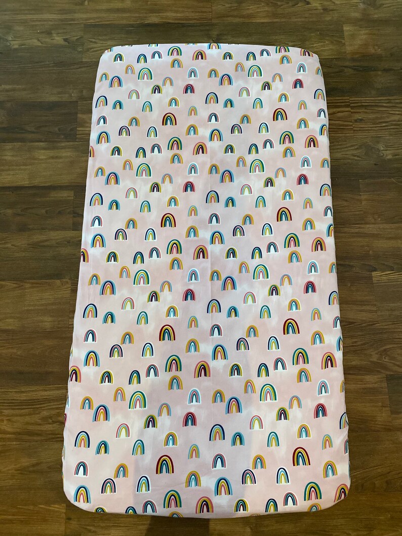 May include: Pink fitted crib sheet with a repeating pattern of colorful rainbows.