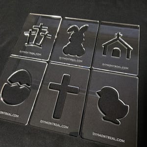 Peut inclure: Ensemble de six pochoirs en acrylique transparent avec différents motifs sur le thème de Pâques, notamment une croix, un lapin, une église, un œuf, un poussin et une fleur. Les pochoirs mesurent tous environ 10 cm sur 10 cm et portent le site Web "DIYMONTREAL.COM" imprimé sur eux.