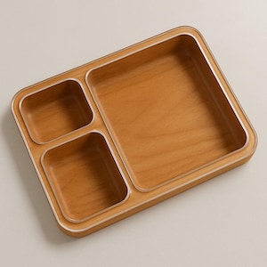 May include: A wooden serving tray with three compartments. The tray is light brown with a silver trim around the edges. The compartments are of different sizes.