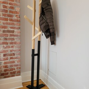 May include: A wooden coat rack with a black base and natural wood branches. A brown jacket is hanging on the rack.
