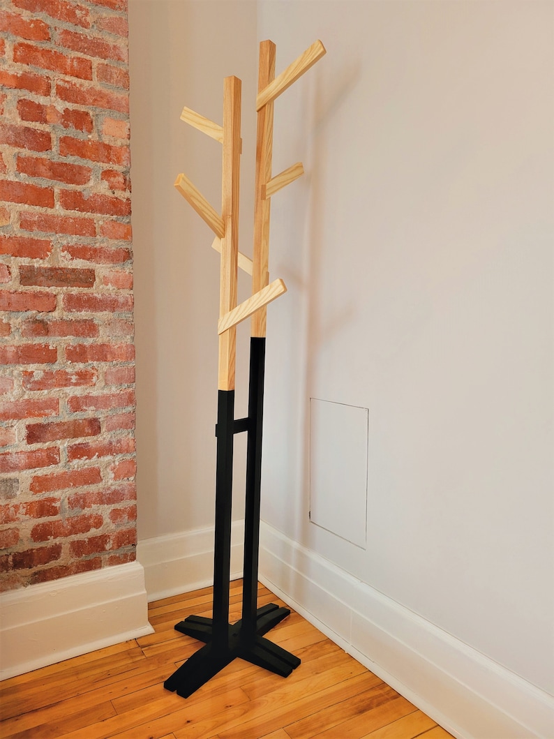 May include: A wooden coat rack with a black base and multiple branches. The rack is standing in a corner of a room with wood floors and white walls.