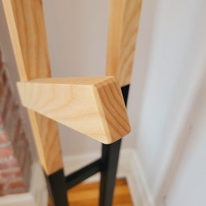 May include: Close-up of a wooden coat rack with a black metal base. The rack has a triangular shape and is made of natural wood.