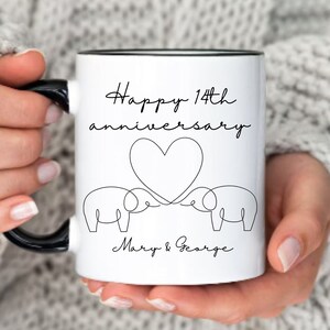 May include: White ceramic mug with a black handle. The mug has a black rim and says "Happy 14th anniversary" with a heart and two elephants. The text at the bottom says "Mary & George".
