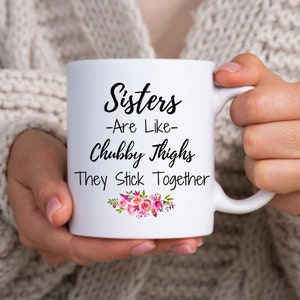 以下が含まれることがあります： 白いセラミック製のマグカップで、黒い文字で「Sisters -Are Like- Chubby Thighs They Stick Together」と書いてあり、底にピンクの花柄のデザインが施されています。