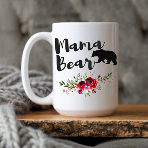 Puede incluir: Taza de caf&eacute; de cer&aacute;mica blanca con una silueta negra de un oso y el texto "Mama Bear" en letras negras. La taza est&aacute; decorada con un dise&ntilde;o floral con flores rojas, rosas y blancas.