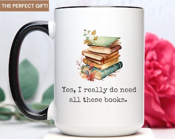 May include: White ceramic mug with a black handle and rim. The mug features a watercolor illustration of a stack of books with autumn leaves and the text "Yes, I really do need all these books."