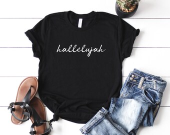 May include: Black t-shirt with the word "hallelujah" written in white cursive lettering. The shirt is laid out on a white wooden surface with a pair of brown sandals, blue jeans, and a potted cactus.
