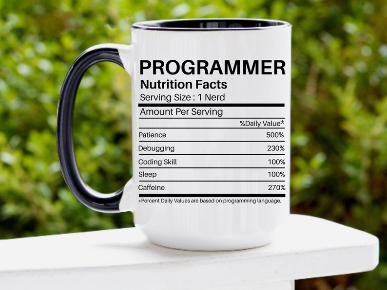 Puede incluir: Taza de cer&aacute;mica blanca con asa y borde negros. La taza presenta la palabra "PROGRAMMER" sobre una etiqueta de "Nutrition Facts", que enumera los valores diarios de Paciencia, Depuraci&oacute;n, Habilidad de Codificaci&oacute;n, Sue&ntilde;o y Cafe&iacute;na. La taza est&aacute; sobre una superficie blanca con fondo verde.