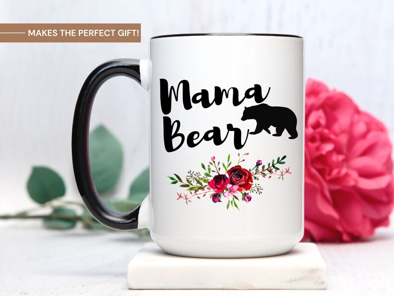 Puede incluir: Taza de cer&aacute;mica blanca con asa y borde negros. La taza tiene una silueta negra de un oso y el texto "Mama Bear" en letra negra. El texto est&aacute; rodeado de un dise&ntilde;o floral con flores rojas, rosas y blancas.