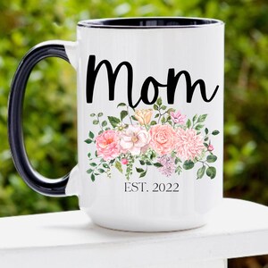 May include: White ceramic mug with a black handle and rim. The mug has a floral design with pink and peach flowers and green leaves. The text "Mom Est. 2022" is printed on the mug.