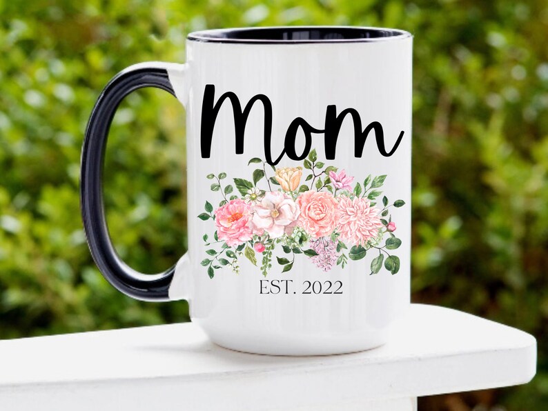 May include: White ceramic mug with a black handle and rim. The mug has a floral design with pink and peach flowers and green leaves. The text "Mom Est. 2022" is printed on the mug.