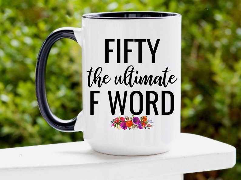 May include: White ceramic coffee mug with a black handle and rim. The mug has black text that reads "FIFTY the ultimate F WORD" with a floral design below the text.