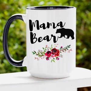 Puede incluir: Taza de cer&aacute;mica blanca con asa y borde negros. La taza tiene una silueta negra de un oso y el texto "Mama Bear" en letras negras. El texto est&aacute; rodeado de un dise&ntilde;o floral con flores rojas y rosas.