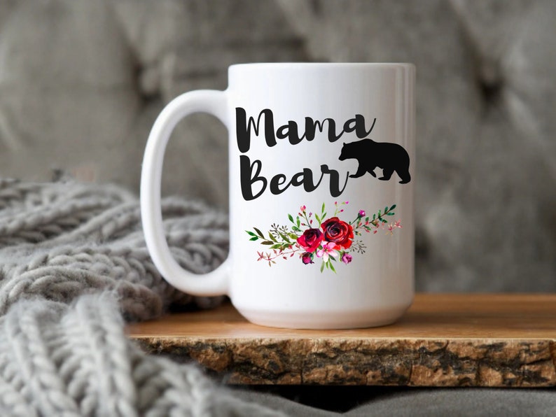 Puede incluir: Taza de caf&eacute; de cer&aacute;mica blanca con una silueta negra de un oso y el texto "Mama Bear" en letras negras. La taza est&aacute; decorada con un dise&ntilde;o floral con flores rojas, rosas y blancas.