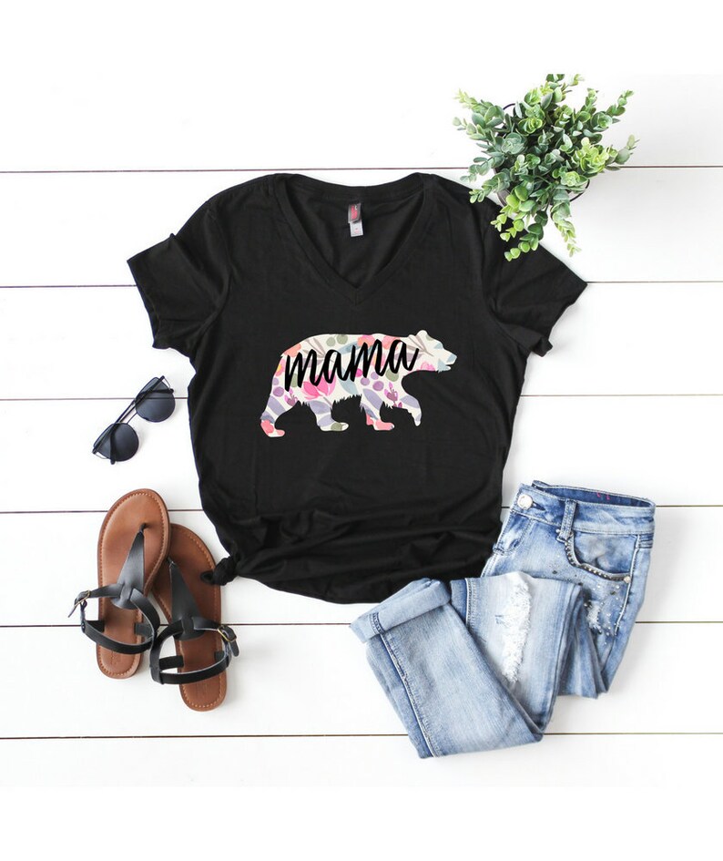 May include: Black v-neck t-shirt with a floral bear silhouette and the word "mama" printed on it. The shirt is laid out on a white wooden surface with a pair of brown sandals, sunglasses, and blue jeans.