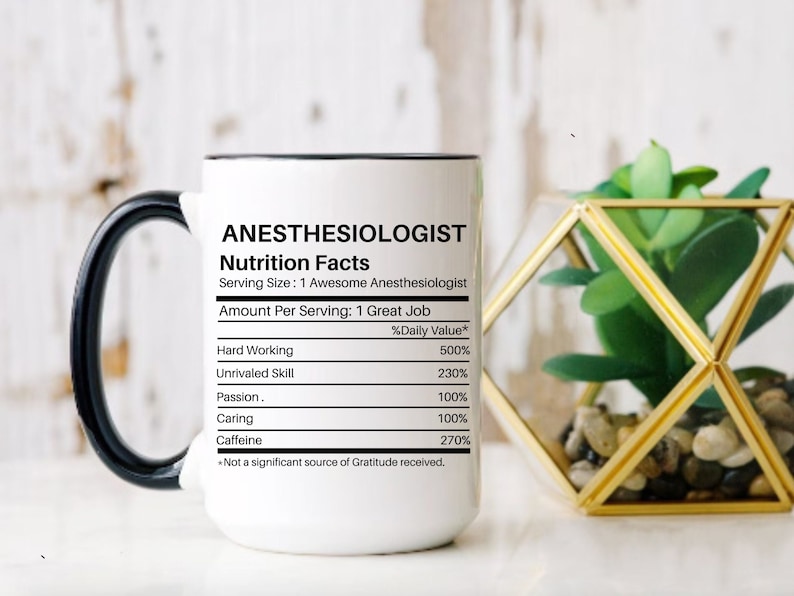 May include: White ceramic mug with a black handle and a black rim. The mug has a nutrition facts label that says "Anesthesiologist" with a list of qualities and their percentage daily value. The mug is sitting on a white surface next to a small gold terrarium with a green plant.