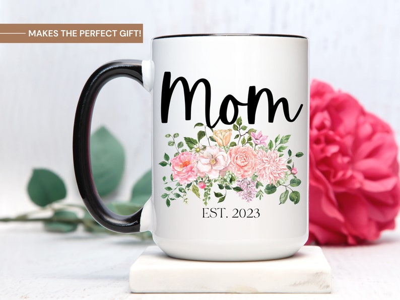 May include: White ceramic mug with a black handle and rim. The mug has a floral design with pink and white flowers and green leaves. The text "Mom Est. 2023" is printed on the mug.