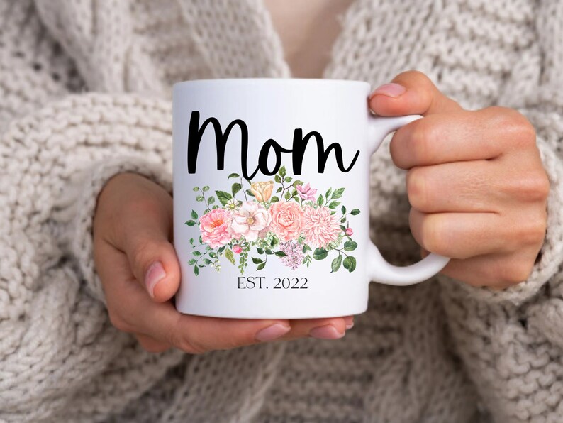 May include: White ceramic mug with a black floral design and the text "Mom Est. 2022".