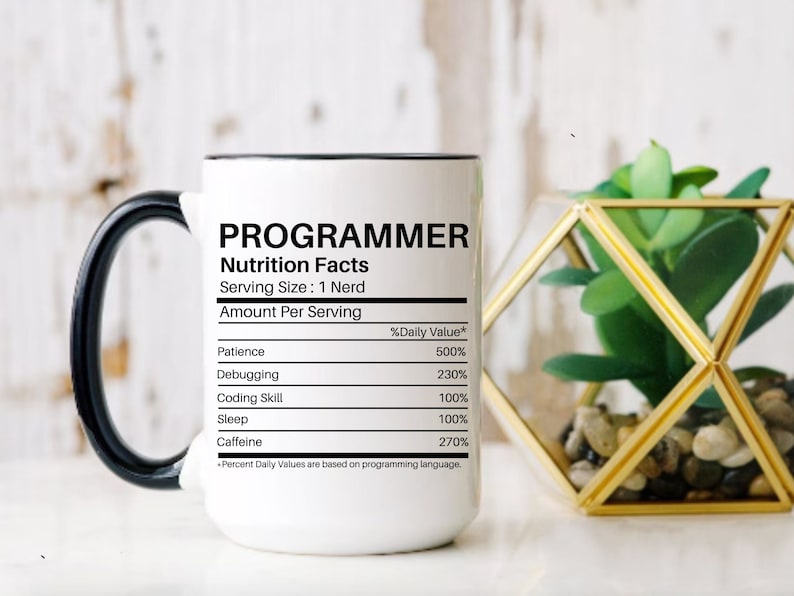 Puede incluir: Taza de cer&aacute;mica blanca con asa y borde negros. La taza presenta la palabra "PROGRAMMER" sobre una etiqueta de "Nutrition Facts". La etiqueta enumera los valores diarios de paciencia, depuraci&oacute;n, habilidad de codificaci&oacute;n, sue&ntilde;o y cafe&iacute;na.