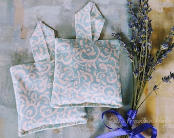 May include: Two small, square, lavender-scented sachets with a light blue and pink floral pattern. The sachets are tied with a blue ribbon and a bunch of lavender is tied with the ribbon. The text "Rocky Mountain Herbs" is visible on the ribbon.