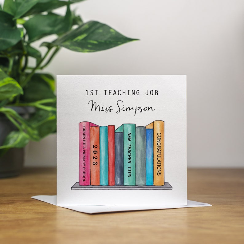 May include: A white card with the text "1st Teaching Job Miss Simpson" and a watercolor illustration of a bookshelf with books titled "Green Hill Primary School", "2023", "New Teacher Tips", and "Congratulations".