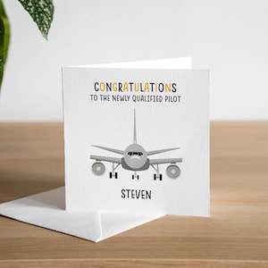 May include: A white card featuring a grey aeroplane illustration. The text reads "CONGRATULATIONS TO THE NEWLY QUALIFIED PILOT" in blue and yellow, with the name "STEVEN" below. The card is on a wooden surface, with a white envelope.
