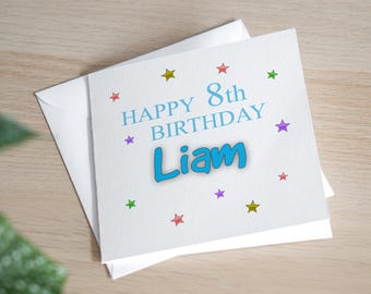 May include: A white birthday card with the text "HAPPY 8th BIRTHDAY Liam" in blue. The card is decorated with colorful stars and comes with a white envelope. The card is on a light wood surface.