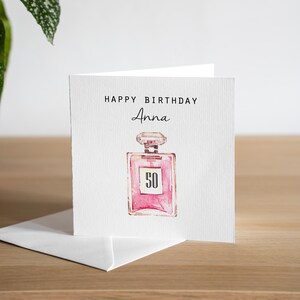 May include: A white greeting card with the text "HAPPY BIRTHDAY Anna" in black. A pink perfume bottle illustration with the number "50" is centered below the text. The card is on a wooden surface.