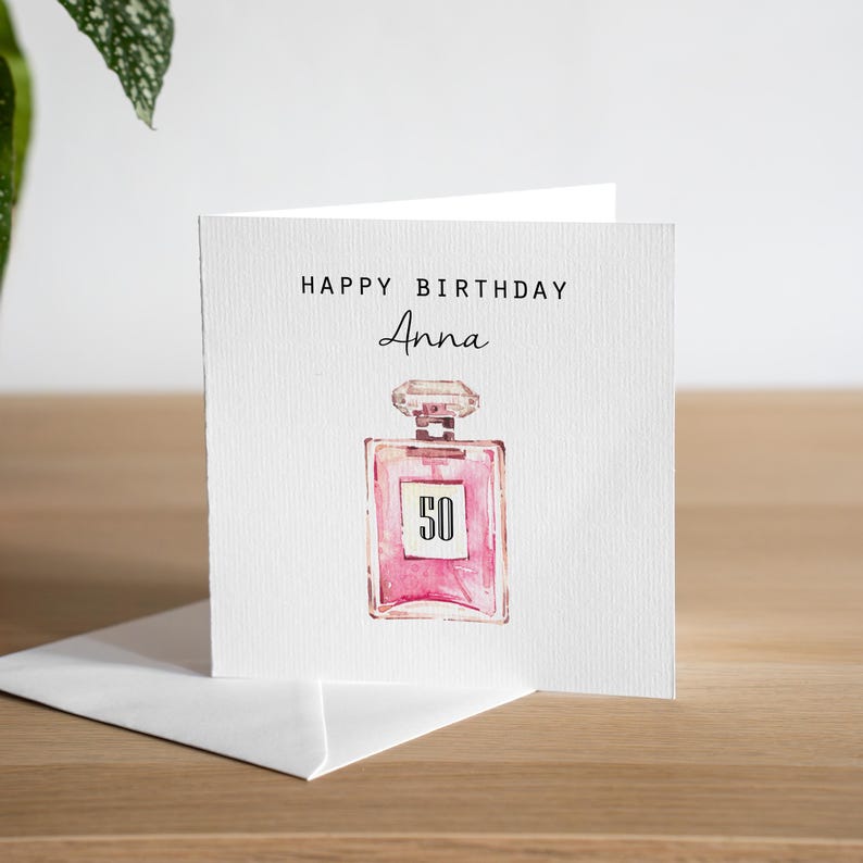 May include: A white greeting card with the text "HAPPY BIRTHDAY Anna" in black. A pink perfume bottle illustration with the number "50" is centered below the text. The card is on a wooden surface.