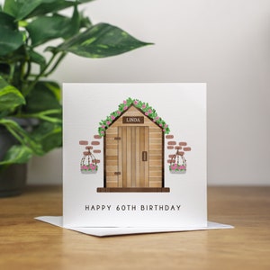 May include: A white card featuring a drawing of a wooden shed with a door and a sign reading "Linda" above it. The shed is adorned with pink flowers and green vines. The text "Happy 60th Birthday" is printed beneath the shed.