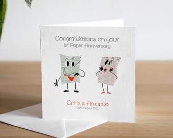 May include: A white card with a cartoon illustration of two pieces of paper with faces. The text on the card reads "Congratulations on your 1st Paper Anniversary" and "Chris & Amanda 30th August 2022".