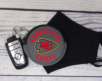 May include: A gray round keychain with the words "Kansas City Chiefs" and a red and yellow arrowhead logo. The keychain is attached to a black face mask.