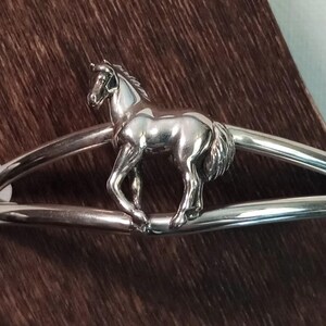 May include: Silver bracelet with a horse design. The horse is in a running pose and is centered on the bracelet.