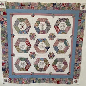May include: A white quilt with a blue and pink border. The quilt is made up of nine hexagonal patches, each with a different floral design. The designs include a watering can, a birdhouse, a wheelbarrow, a teacup, a heart, and a bouquet of flowers.