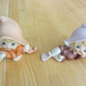 Puede incluir: Dos figuritas de cerámica de elfos con sombreros puntiagudos. Un elfo lleva un atuendo marrón y naranja y está sentado. El otro elfo lleva un atuendo marrón y morado y está acostado. Ambos elfos tienen el pelo rubio y ojos grandes.