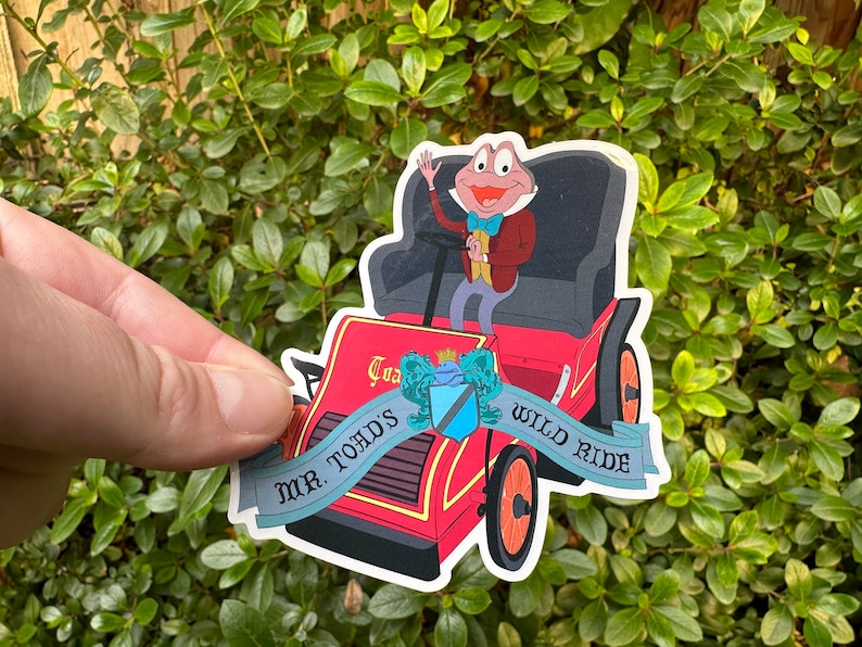 May include: A red and blue sticker featuring Mr. Toad's Wild Ride from the Disney theme park. The sticker shows Mr. Toad driving a red car with a blue banner that reads "Mr. Toad's Wild Ride".