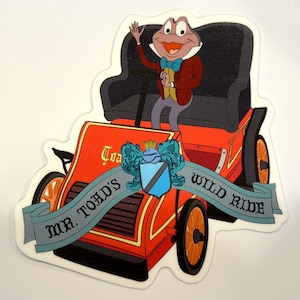 May include: A red and black cartoon car with a green frog wearing a suit and a bow tie driving. The car has a banner that says "Mr. Toad's Wild Ride".