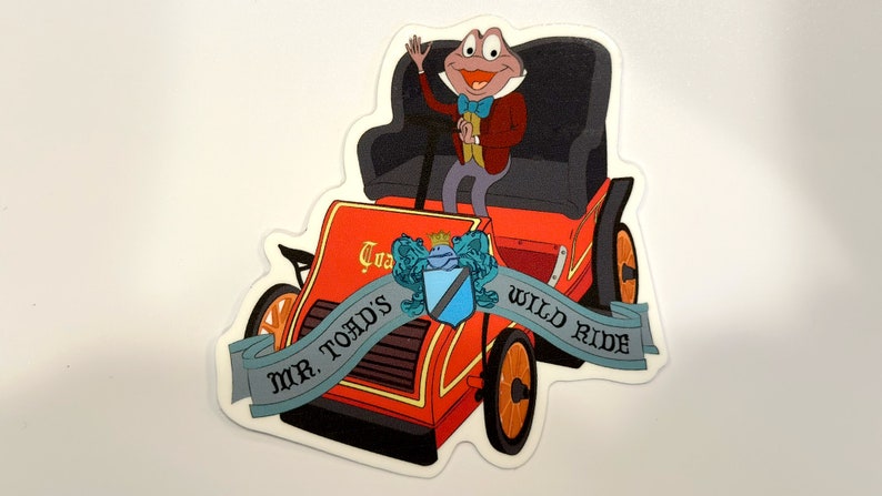 May include: A red and black cartoon car with a green frog wearing a suit and a bow tie driving. The car has a banner that says "Mr. Toad's Wild Ride".