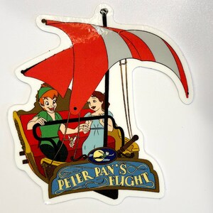 May include: A white sticker featuring a red and white striped sail, Peter Pan and Wendy flying in a red and gold ship. The text on the sticker reads "Peter Pan's Flight".