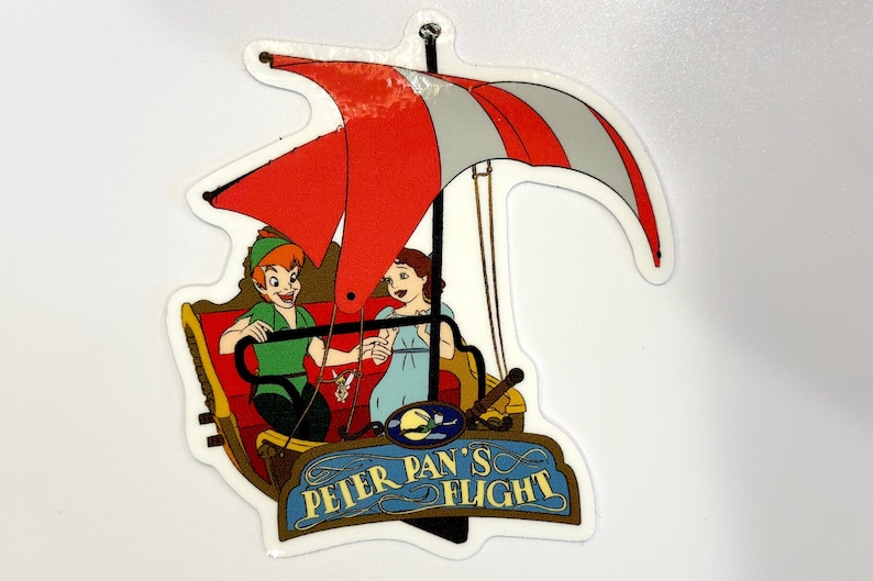 May include: A white sticker featuring a red and white striped sail, Peter Pan and Wendy flying in a red and gold ship. The text on the sticker reads "Peter Pan's Flight".