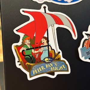 May include: A red, white and blue Peter Pan's Flight ride sticker with Peter Pan and Wendy flying in a boat with a red and white sail. The sticker has a blue sign that says "Peter Pan's Flight".