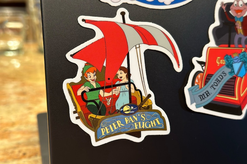 May include: A red, white and blue Peter Pan's Flight ride sticker with Peter Pan and Wendy flying in a boat with a red and white sail. The sticker has a blue sign that says "Peter Pan's Flight".