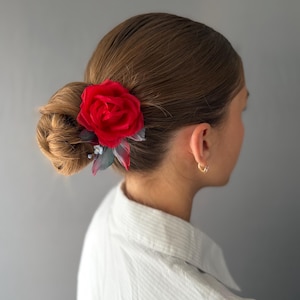 Pode incluir: Um acessório de cabelo com uma grande rosa de tecido vermelho vibrante com pétalas em camadas, acentuada por folhas menores cinza-escuras e pequenos botões de flores brancas, presa num coque castanho.