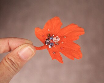 May include: A close-up of an orange felt maple leaf with a cluster of small, colorful rhinestones in the center.