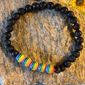 Può includere: Braccialetto con perline di pietra lavica nera e sei perline a righe arcobaleno. Il braccialetto è elastico e poggia su una pietra strutturata marrone chiaro. Un simbolo di orgoglio.