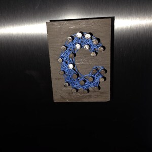 May include: A wooden plaque with the letter "C" crafted from blue string and nails. The letter is centered on a rectangular, gray-stained wooden base. The nails are silver and evenly spaced, creating the letter's form.
