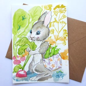 May include: Watercolor illustration of a gray bunny holding a radish and a bag of peas. The bunny has blue eyes and is surrounded by green and yellow foliage. The bag has a floral pattern. The artwork is on a white card with a brown envelope.