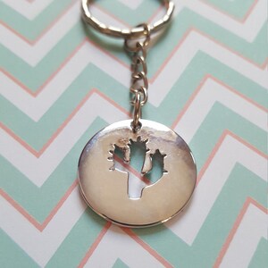 Könnte beinhalten: Silberner Schlüsselanhänger mit einem runden Anhänger mit einem ausgeschnittenen Kaktus-Design. Der Anhänger ist an einer silbernen Kette und einem silbernen Schlüsselring befestigt.