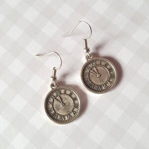 Può includere: Orecchini in argento a forma di quadrante di orologio con numeri romani. Le lancette dell'orologio sono quasi alle 12.