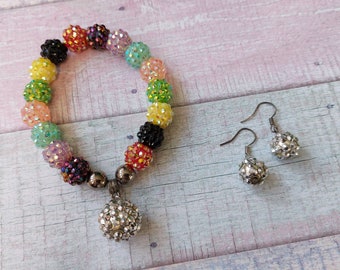 Op de afbeelding: Een kleurrijke kralen armband met een zilveren bedel en bijpassende zilveren oorbellen. De armband is gemaakt van kleine, ronde kralen in tinten roze, groen, geel, rood en zwart. De oorbellen zijn eenvoudige zilveren hoepels met een zilveren bolvormige bedel.